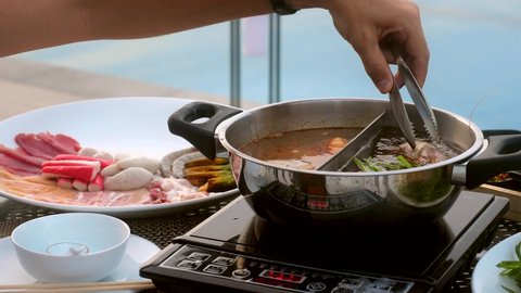 Shabu shabu or sukiyaki meat, beef and seafood in boiling hot pot dish near poolside Japanese restaurant. Man hand put a big shrimp in shabu shoyu and spicy base in hotpot. Close up
