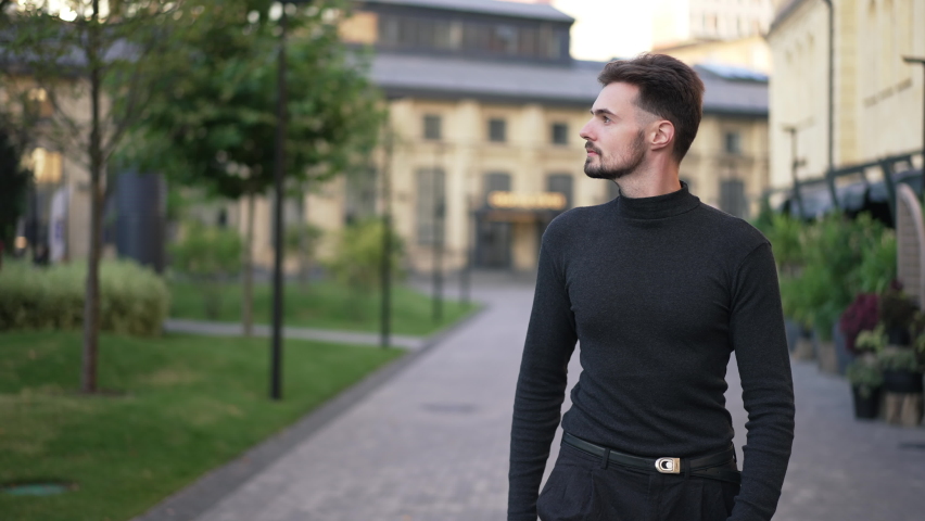 Front view portrait of young Caucasian handsome man walking in city looking around. Confident brunette bearded guy strolling outdoors in slow motion in the morning. Lifestyle concept