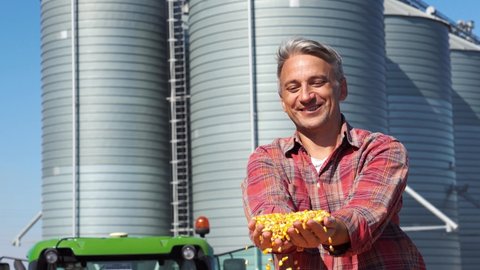 Happy Farmer Showing Freshly Harvested Grains In His Hands Against Grain Storage Silo. Farmer Sitting In Tractor Trailer Full Of Corn Seeds In front Of Grain Silos. Ethanol Biorefinery Silos.