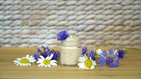  Natural homemade eye cream in transparent glass jar with cornflowers and chamomile flowers on the beige, white background.                            
