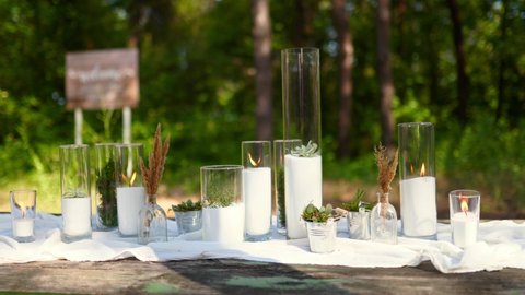 Wedding party banquet outdoors in pine forest. Dining tables decorated in boho style with candles in glass vases, white cloth, succulent flowers. Eco organic floral composition decor for holiday.