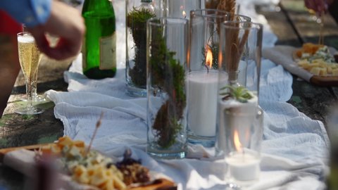 Person takes cheese from dining table decorated in boho style with candles, cloth, flowers, served with tableware, dishes, meals, stemware, drinks. Wedding party banquet catering outdoors in forest.