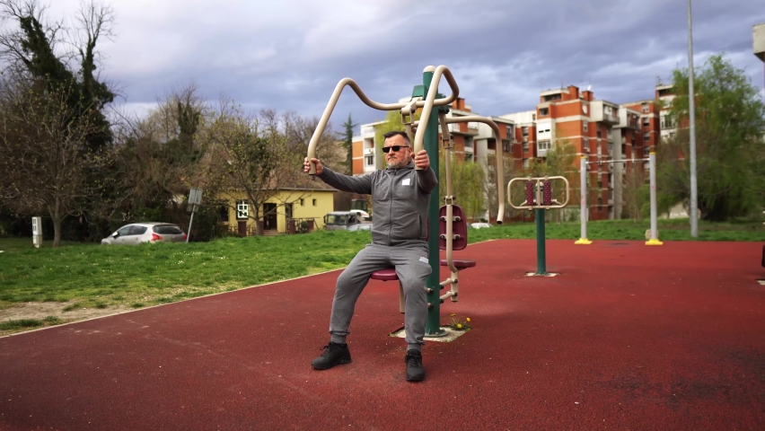 An elderly man with a gray beard and sunglasses, in sportswear, is doing arm exercises on a gym machine, in an outdoor gym.
