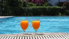 Two bright orange glasses with cocktail or juice with ice stand edge of pool, blue water, against backdrop of beautiful green, summer day. Summer pool party. Slow motion video. Vacation, detox.