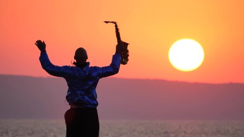 Young slender musician saxophonist with golden alt saxophone dances and plays against backdrop of sunset, big orange sun, dawn and sea. The concept of romantic music. Slow motion video. Saxophonist.