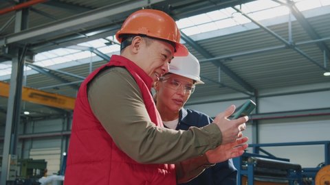 Happy two factory technicians with helmet use mobile phone standing in factory. Woman and asian man. Engineers, team