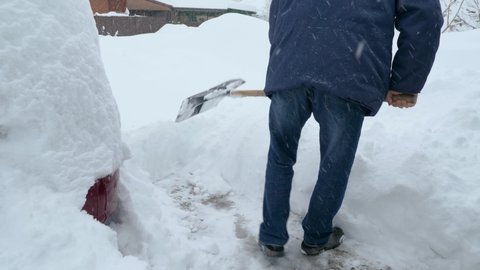View from behind a man in a jacket and jeans is shoveling snow next to his house. Clearing the area in winter after a snow storm. Snowfall in the background