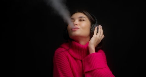 Beautiful young woman in a pink sweater listens to music with wireless headphones, dances and smokes an electronic cigarette. Vaping smoke on a black background