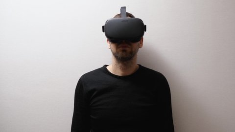 A bearded man in a black sweatshirt standing against a gray wall removes VR helmets from your head and puts them on you. VR glasses for entertainment, learning, and gaming