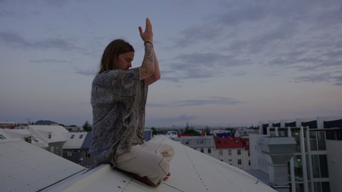 Slow Motion Wide Shot Of Young Man With Long Hair And Tattoos Sitting Cross Legged With Bare Feet On Rooftop, Meditating And Stretching In Reykjavik, Iceland