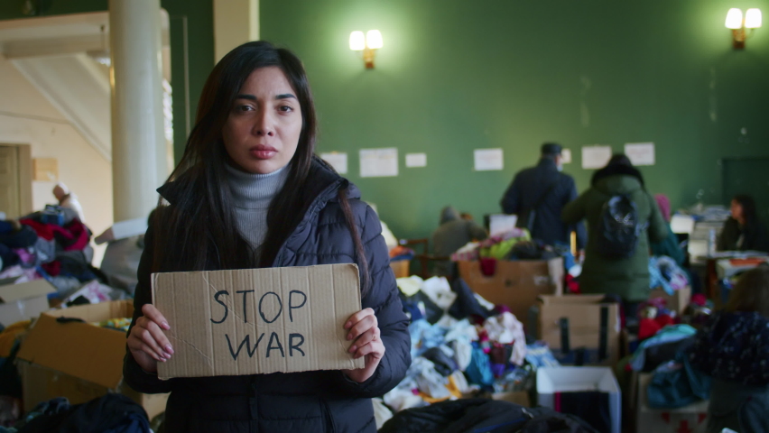 A portrait of a sad Armenian woman in the large hall of the refugee assistance center among a large number of clothes and refugees. War between Russia and Ukraine. Royalty-Free Stock Footage #1089019393