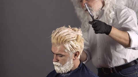 Hair coloring man. Crazy fun hairdresser. Making of a dyed hair for a bearded hipster guy. Beard coloring man agains grey hair. Bearded man, barber shop.