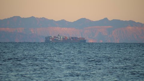 Cargo ship transporting goods off coast of Sinai. Freighter carries shipping containers near shore of Saudi Arabia. Merchant vessel, watercraft moving past rocky place. Shipment, reaching load by sea