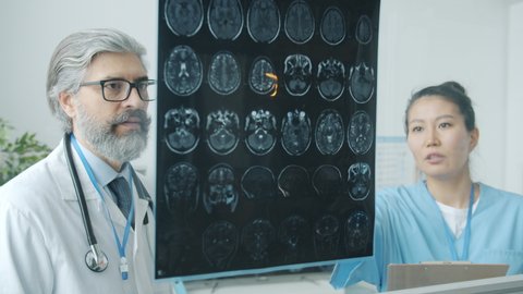 Neurologist man in uniform analyzing MRI brain scans and talking to Asian nurse in modern clinic. Professional occupation and medicine concept.