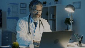 Mature male doctor in white coat working with laptop and drinking coffee concentrated on job in dark office at night. Pandemic and medicine concept. - Powered by Shutterstock - Get 15% off with code: PIKWIZARD15