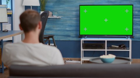 Static tripod shot of man switching channels while looking at green screen on tv and sitting on sofa. Over shoulder view of person relaxing using television remote zapping on chroma key display.