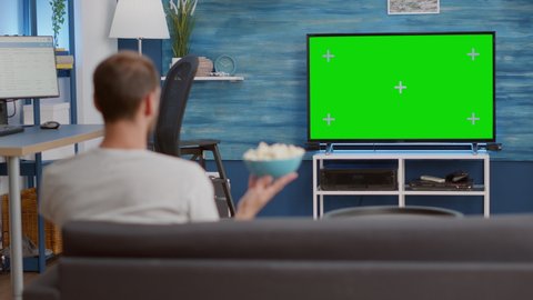 Man eating popcorn from bowl while watching favourite tv show on green screen while sitting comfortable on couch. Person sitting on sofa looking at television show on chroma key display in living room