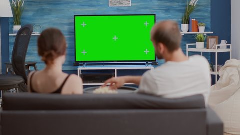 Static tripod shot of couple having a snack while looking at green screen tv watching a movie and turning up the volume. Woman and man watching chroma key television and zapping while sharing popcorn.