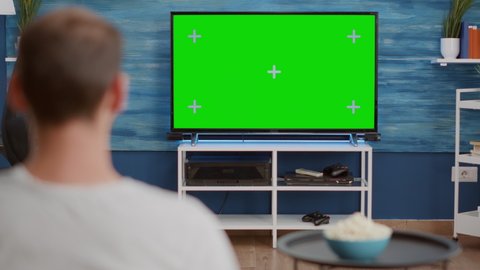 Over shoulder view of man switching channels while looking at green screen on tv and sitting on sofa in modern living room. Person relaxing using television remote zapping on chroma key display.