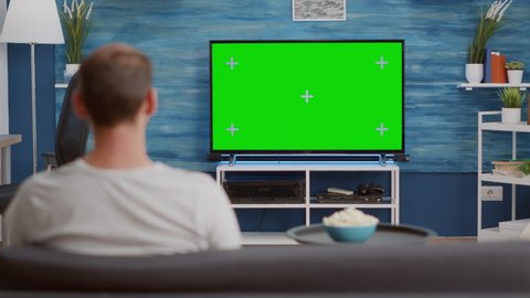 Man sitting on sofa looking at green screen on tv relaxing watching movie in modern living room with bowl of popcorn. Back view of person sitting on couch in front of television with chroma key