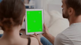 Closeup of couple holding vertical digital tablet with green screen in online conference or group video call in home living room. Man and woman talking in front of touchscreen device with chroma key.