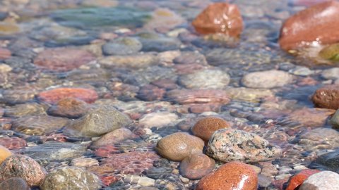 colorful pebble stones in water in sunny day, sea ocean waves gently moving among big pebbles. rocky ground in water