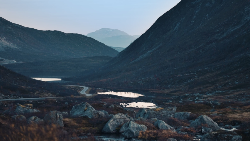 A narrow road snaking through the vast valley. Mountains tower on both sides. lakes and puddles scattered through the valley. Aerial view.