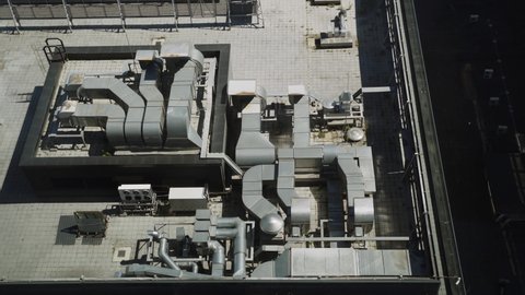 Serviced roof of industrial building is covered paving slabs with ventilation system with many metal air ducts of various sizes and air conditioners with growing green plants between tiles. Top view.