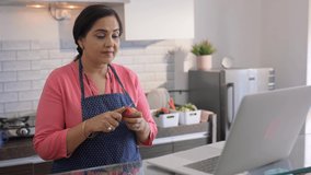 Indian Asian middle aged Woman or Female wearing chef apron standing in front of a laptop in modern kitchen house setup chopping tomato while taking online cooking Tutorials courses to prepare recipe.