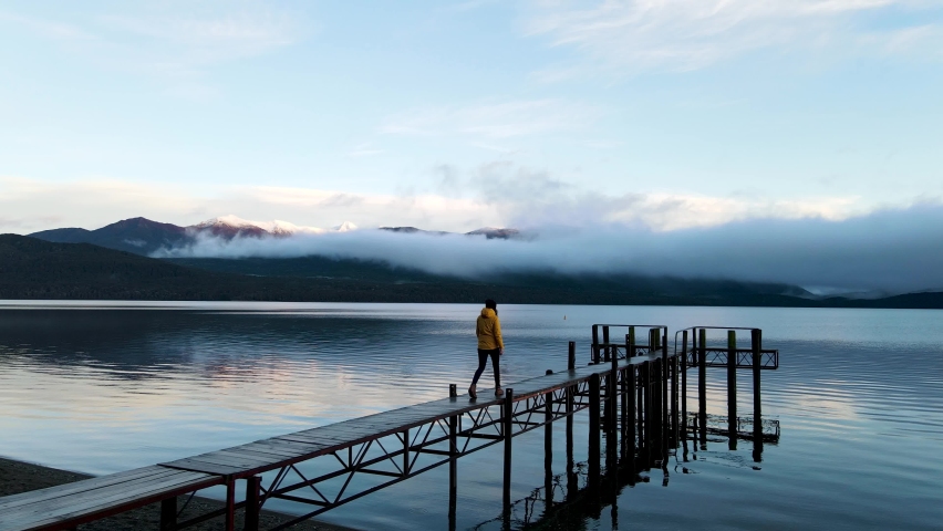 Female walking on a wooden pier at a lake at sunset. Woman with a yellow jacket walking on a duck pond enjoying the beautiful view of the mountains in New Zealand while a drone orbits around.