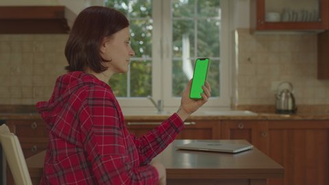 Young adult woman using mobile phone green screen vertically sitting in the kitchen. Female touching vertical cell phone chroma key.