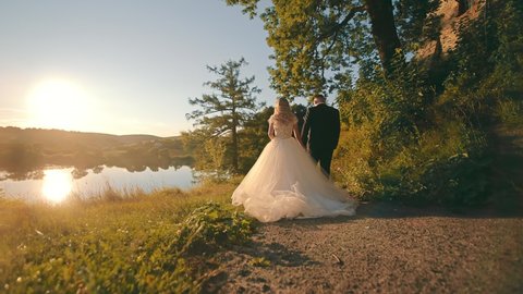 Happy bride in white dress, and groom walking down country road, holding hands in setting sun. Back view of wedding couple walking, with scenic lake and balloons on background. Concept of wedding