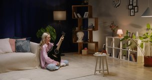 Woman playing acoustic guitar and recording video on smartphone camera in living room, video lesson. Female blogger making content for channel. Lifestyle vlog, talking at camera webcam online.