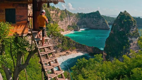 Bali, Nusa Penida - Indonesia - Jan 20 2022: Travel people standing near scenery wooden hut and enjoy amazing top view on coastline Diamond beach Bali, Indonesia 4K Aerial view