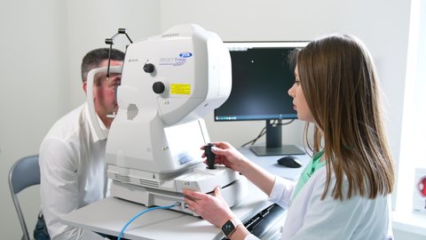 KYIV, UKRAINE - August 2021: Long-haired young female oculist sitting at apparatus examining man's eyesight. Doctor moves the device lever arm slowly.