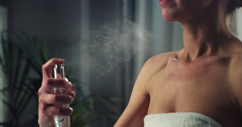 Cinematic close up of young woman in white bathrobe is applying luxurious female scent fragrant perfume on her neck after shower during personal beauty hygiene routine at home before going out.