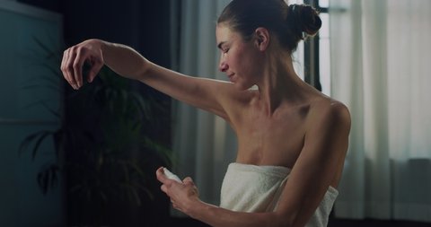 Cinematic shot of young woman in white bathrobe is applying female antiperspirant body spray deodorant on her armpit after shower during personal beauty hygiene routine at home before going out.