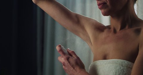 Cinematic close up of young woman in white bathrobe is applying female antiperspirant body spray deodorant on her armpit after shower during personal beauty hygiene routine at home before going out.