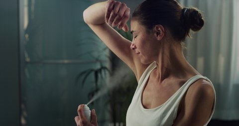 Cinematic close up of young woman applying female antiperspirant body dry spray deodorant on armpit and looking in mirror during morning beauty and personal hygiene routine at home before going out.