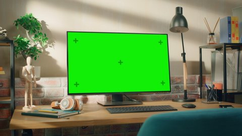 Desktop Computer Monitor Standing on a Table with a Green Screen Chromakey Mock Up Display. Cozy Empty Loft Apartment with a Lamp, Notebooks and Smartphone on the Table. Arc Shot.
