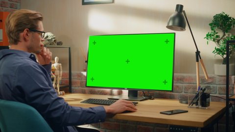 Young Handsome Man Working from Home on Desktop Computer with Green Screen Mock Up Display. Creative Male Checking Social Media, Browsing Internet. Living Room in Bright Loft Apartment. Arc Shot.