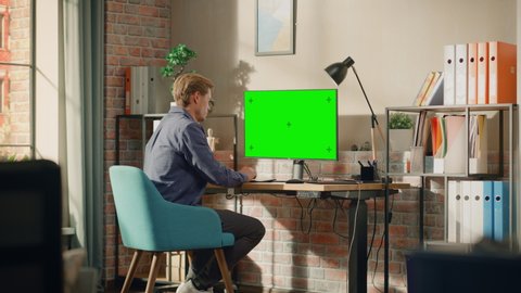 Young Handsome Man Working from Home on Desktop Computer with Green Screen Mock Up Display. Male Checking Corporate Accounts, Messaging Colleagues. Loft Living Room with Big Window. Zoom In Shot.