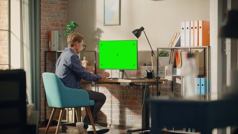 Young Handsome Man Working from Home on Desktop Computer with Green Screen Mock Up Display. Male Checking Corporate Accounts, Messaging Colleagues. Loft Living Room with Big Window. Wide Shot.