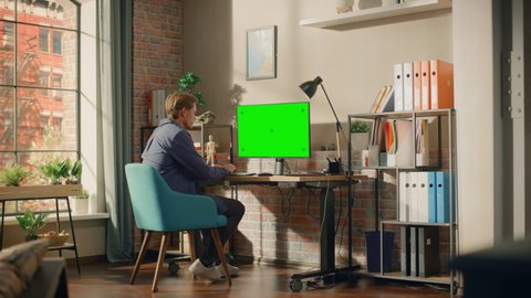 Young Handsome Man Sitting Down to Work from Home on Desktop Computer with Green Screen Mock Up Display. Creative Male Checking Social Media, Browsing Internet. Living Room in Bright Loft Apartment.