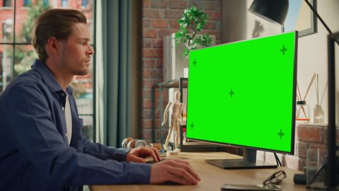 Young Handsome Man Working from Home on Desktop Computer with Green Screen Mock Up Display. Creative Male Checking Social Media, Browsing Internet. Living Room in Bright Loft Apartment. Static Shot.