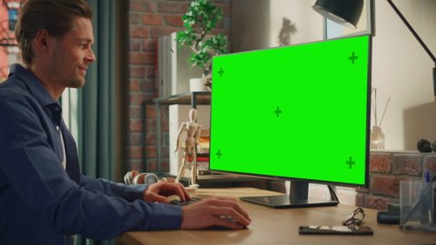 Young Handsome Man Working from Home on Desktop Computer with Green Screen Mock Up Display. Male Checking Corporate Accounts, Messaging Colleagues. Loft Living Room with Big Window. Arc Shot.