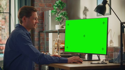 Young Handsome Man Working from Home on Desktop Computer with Green Screen Mock Up Display. Male Checking Corporate Accounts, Messaging Colleagues. Loft Living Room with Big Window. Zoom In Shot.