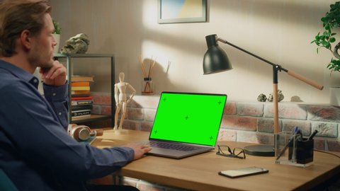 Young Handsome Man Working from Home on Laptop Computer with Green Screen Mock Up Display. Creative Male Checking Social Media, Browsing Internet. Living Room in Loft City Apartment.