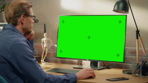 Young Handsome Man Working from Home on Desktop Computer with Green Screen Mock Up Display. Creative Male Checking Social Media, Browsing Internet. Living Room in Bright Loft Apartment. Arc Shot.