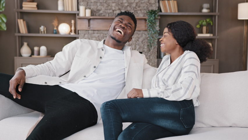 Happy african american couple jumping falls on soft sofa comfortable couch laugh enjoy relax new furniture real estate relocation rest celebrate move day to own home rented flat. Homeowners family Royalty-Free Stock Footage #1089056993